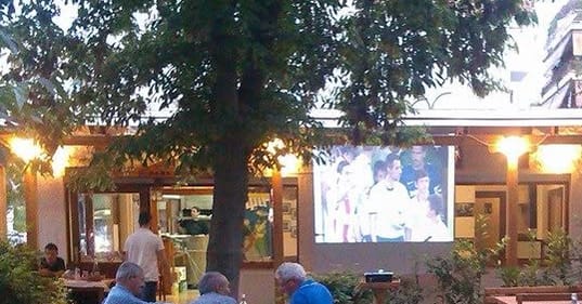 Ζεστά φώτα κάτω από την ξύλινη πέργκολα και τα δέντρα του χώρου, το σούρουπο.